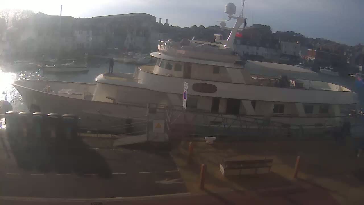 A large motor yacht is docked at a harbor on a hazy, sunny day, with smaller boats visible in the background.