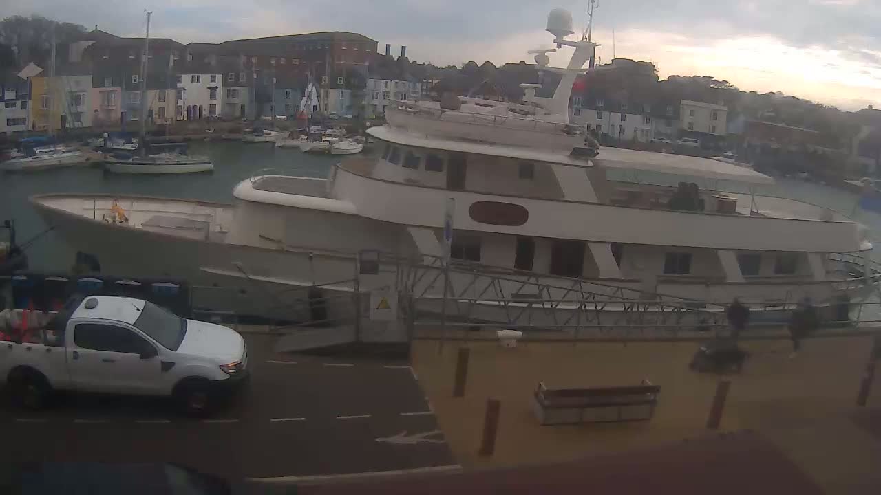 A large motor yacht is docked in a harbor next to a white pickup truck, under an overcast sky.