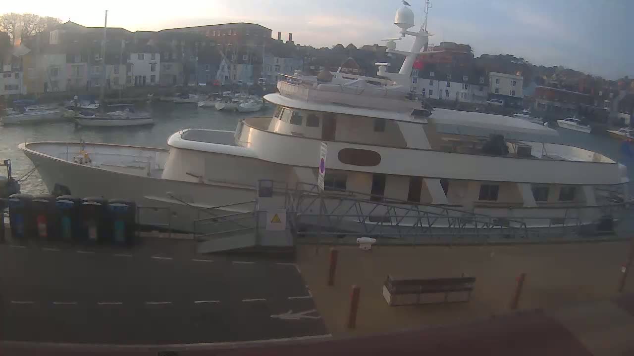 A large, white motor yacht is docked at a harbor filled with smaller boats, under a partly cloudy sky.