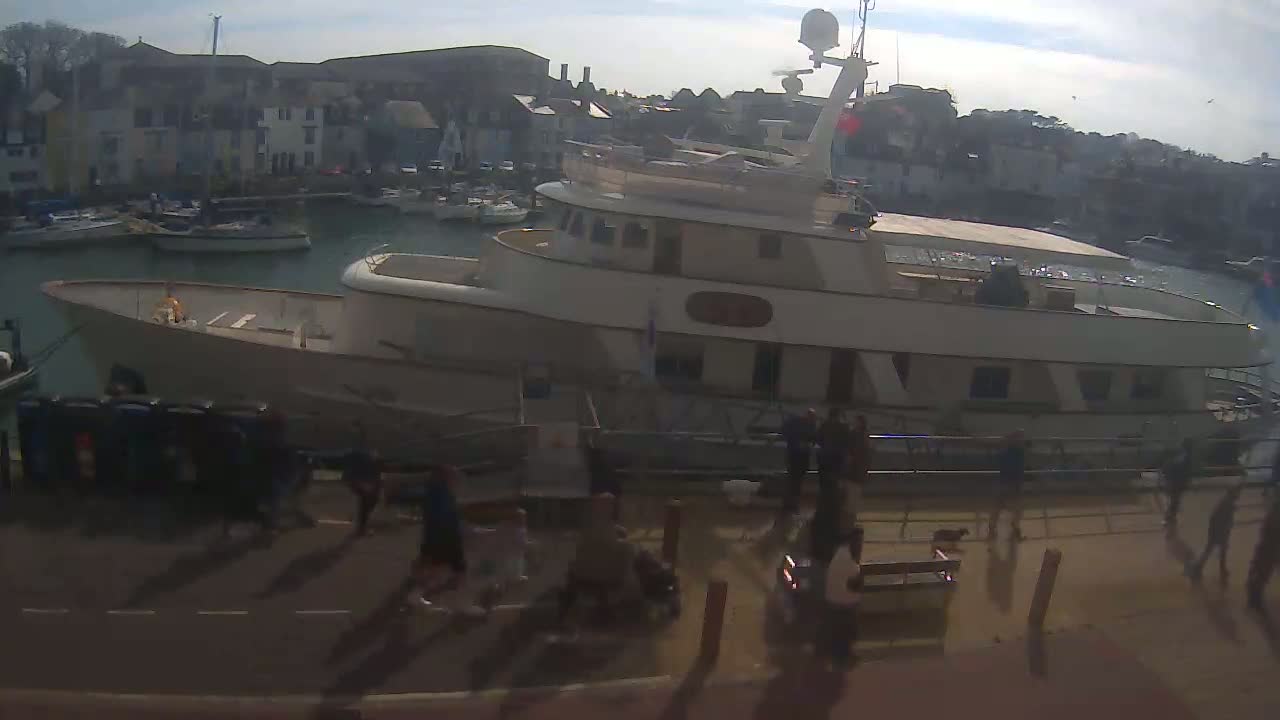 A large, light-colored motor yacht is docked in a harbor on a sunny day, with several people walking on the dock near it and smaller boats in the background.