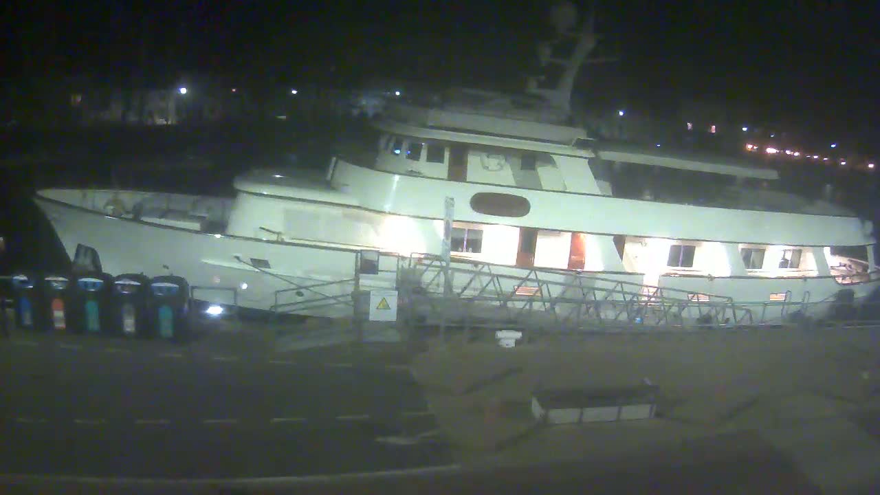 A large, white motor yacht is docked at night, illuminated by interior lights, with a gangway extending to the shore.