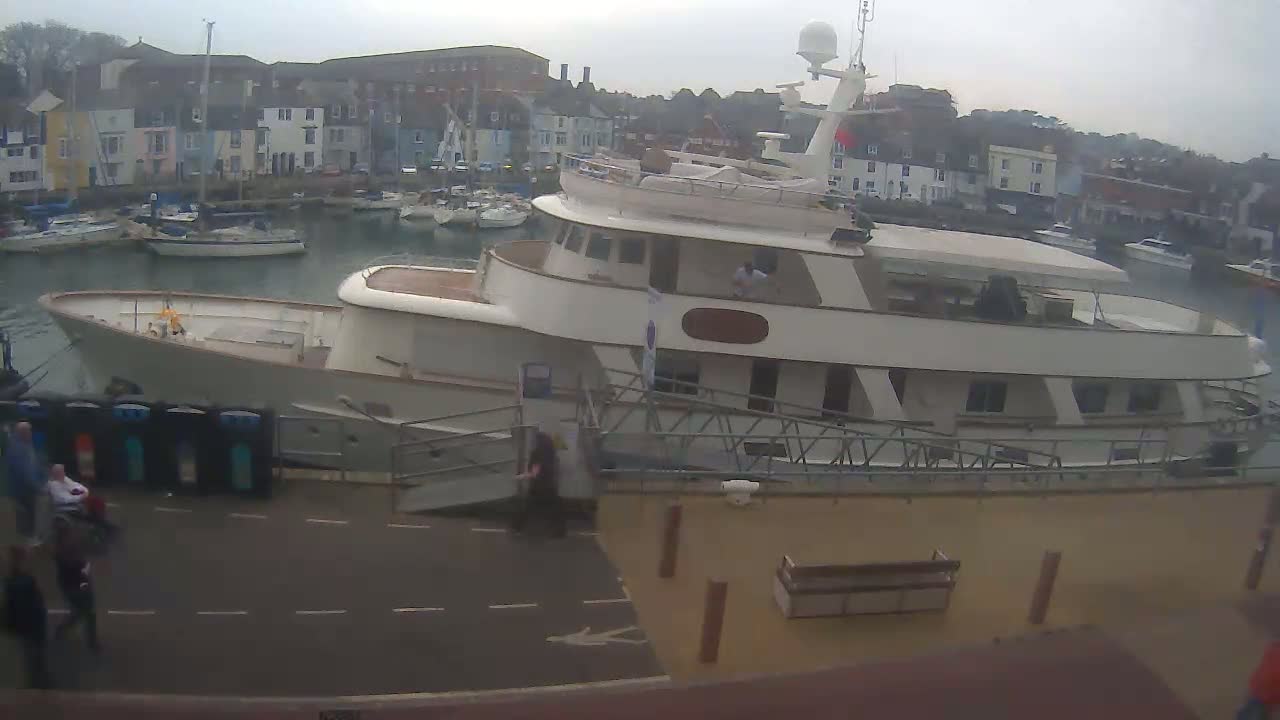 A large, light-colored motor yacht is docked at a marina on an overcast day, with several smaller boats visible in the background.