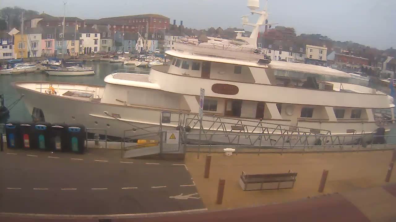 A large, light-colored motor yacht is docked in a harbor on an overcast day, with numerous smaller sailboats visible in the background.