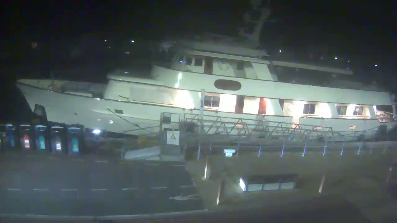 A large, white motor yacht is docked at night, illuminated by interior lights, next to a metal gangway.
