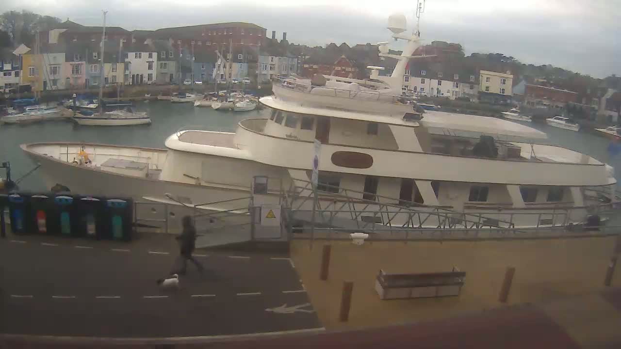 A large motor yacht is docked in a harbor filled with smaller boats, under an overcast sky.