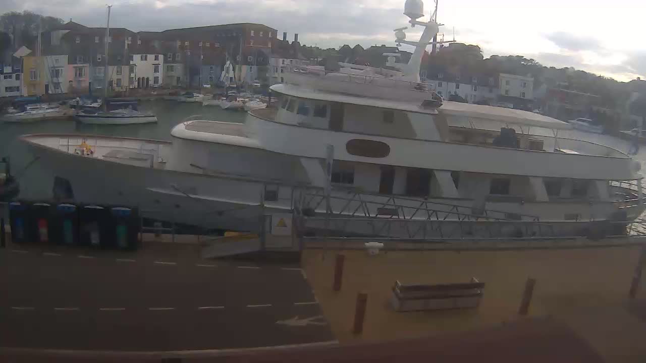 A large motor yacht is docked in a harbor, with overcast skies and several smaller boats visible in the background.