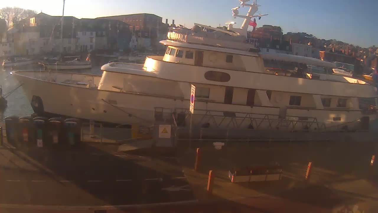 A large, light-colored yacht is docked at a harbor at sunset, with other smaller boats visible in the background.