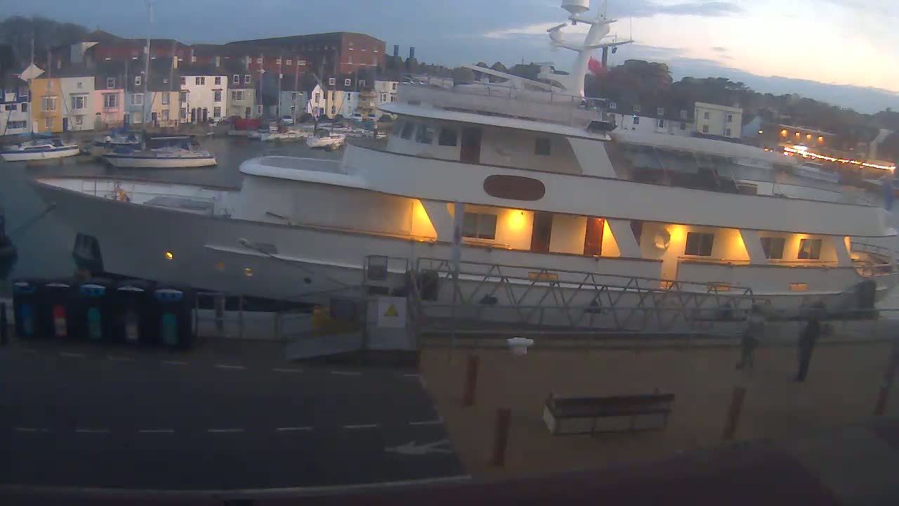 A large, white yacht is docked at a harbor at dusk, with smaller boats visible in the background.