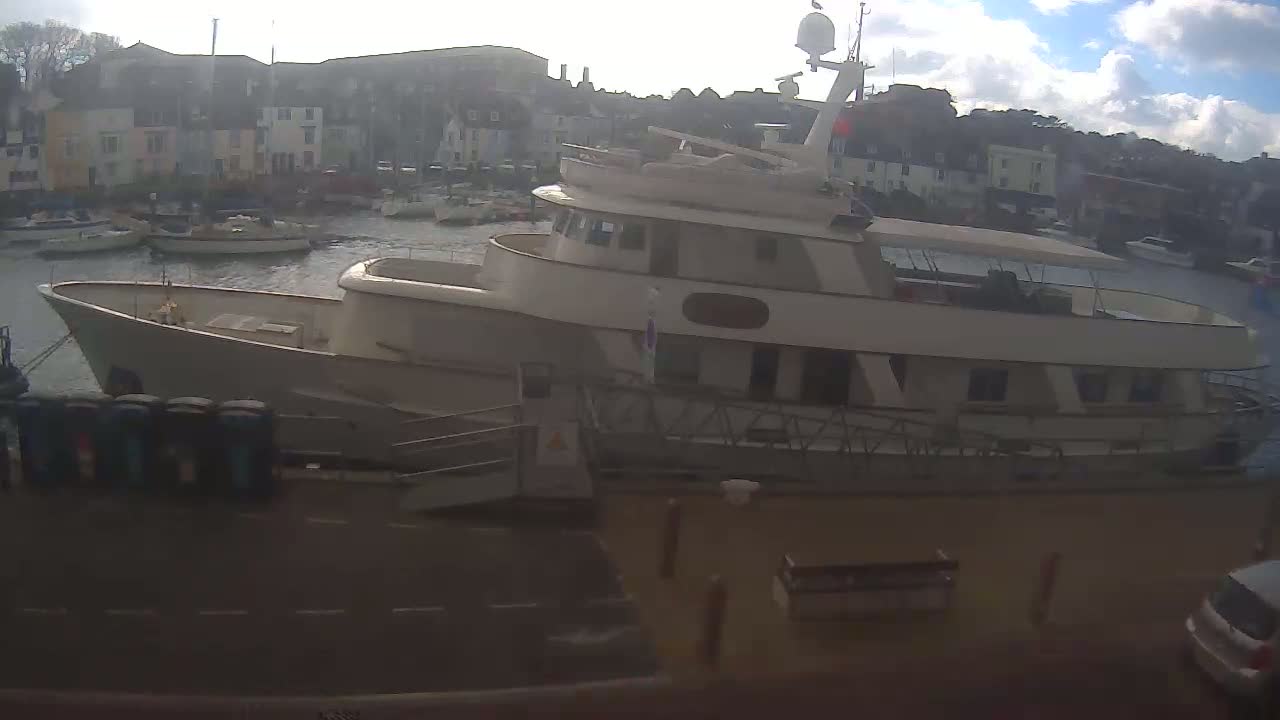 A large, light-beige motor yacht is docked in a harbor on a partly sunny day with some clouds.