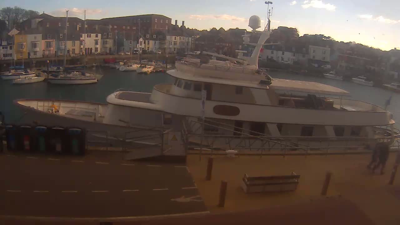 A large motor yacht is docked in a harbor with several smaller sailboats, under a partly cloudy sky.