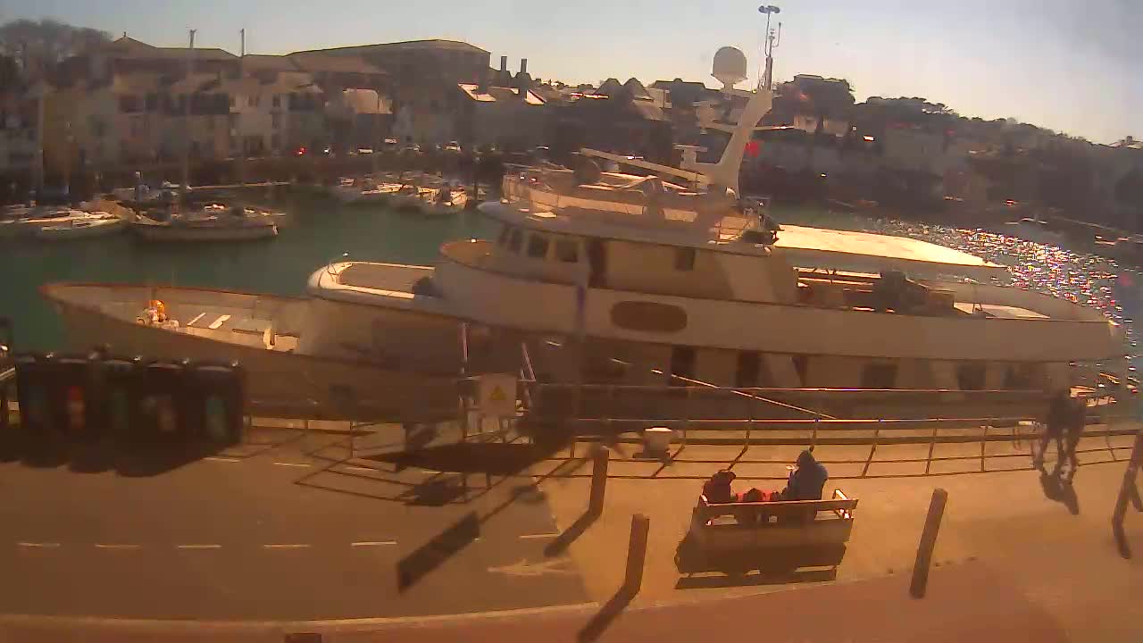 A large, light-colored motor yacht is docked at a harbor on a sunny day, with several smaller boats in the background and people near the dock.