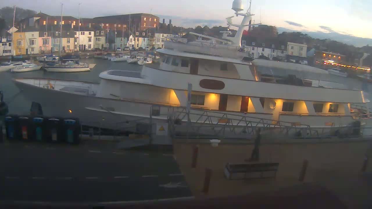 A large motor yacht is docked at a harbor in the evening, with many smaller boats in the background under a twilight sky.