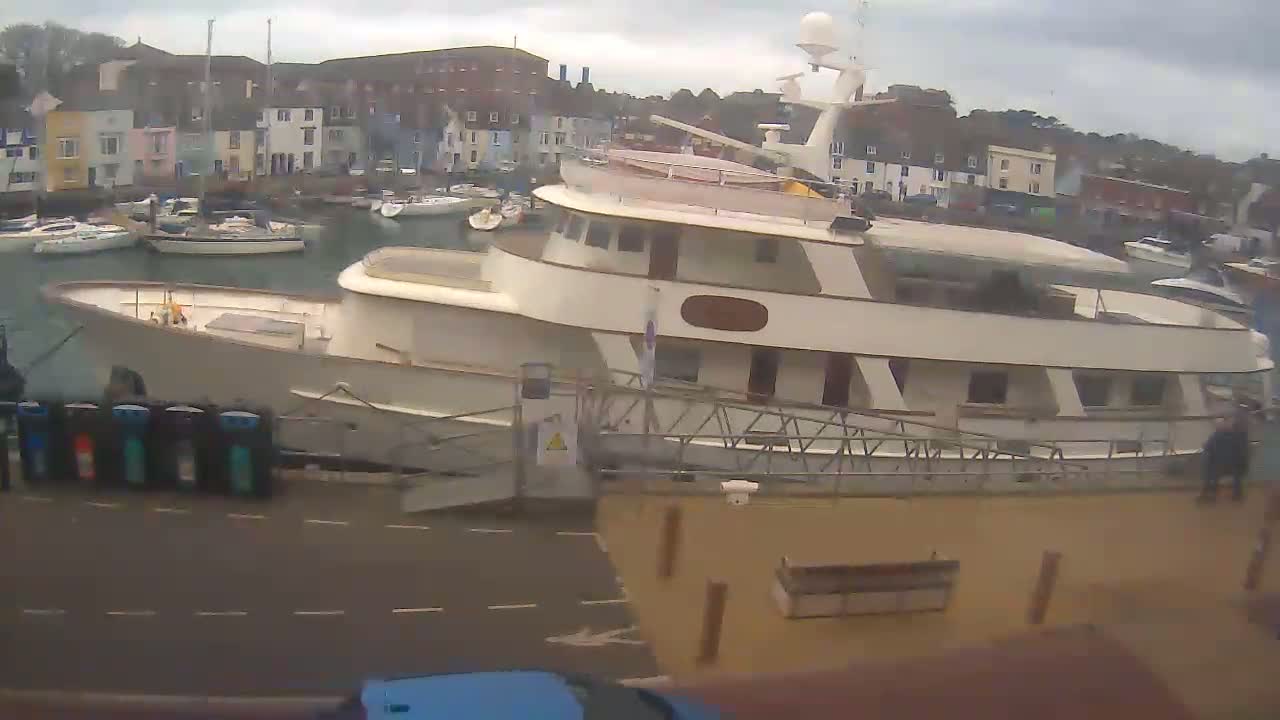 A large, light-colored motor yacht is docked at a harbor filled with smaller boats, under an overcast sky.