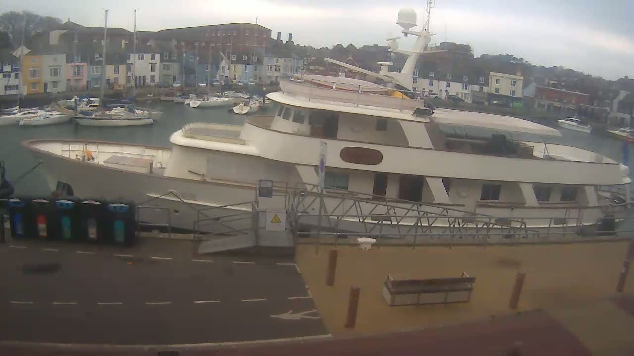 A large motor yacht is docked at a marina on an overcast day, with several smaller sailboats and motorboats visible in the background.