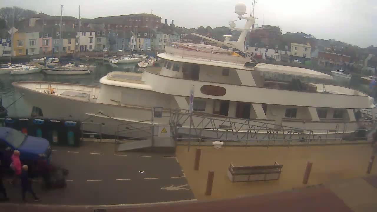 A large motor yacht is docked at a harbor on an overcast day, with several smaller sailboats visible in the background and a few people walking near the vessel.