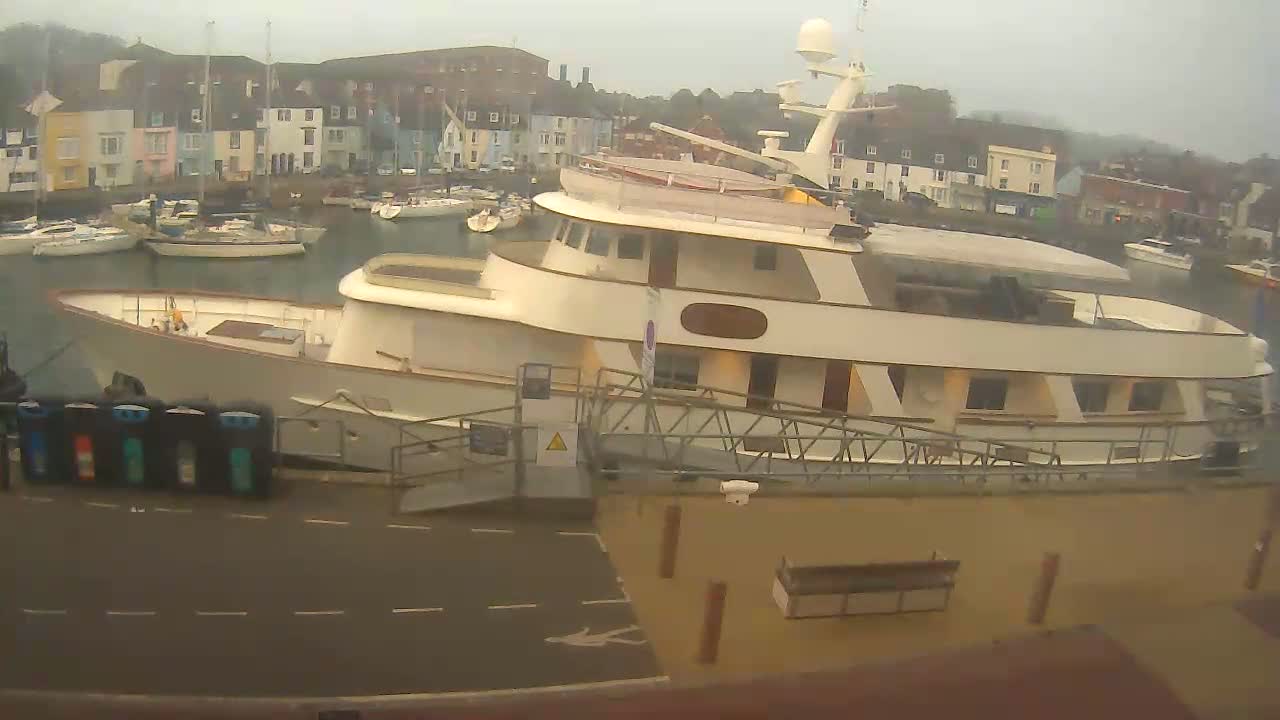 A large, light-colored motor yacht is docked at a harbor in overcast weather, with smaller sailboats visible in the background.