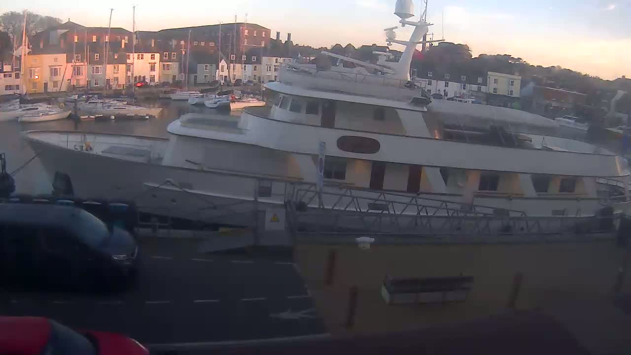 A large motor yacht is docked in a harbor at sunset, with numerous smaller boats in the background and cars visible on a road in the foreground.