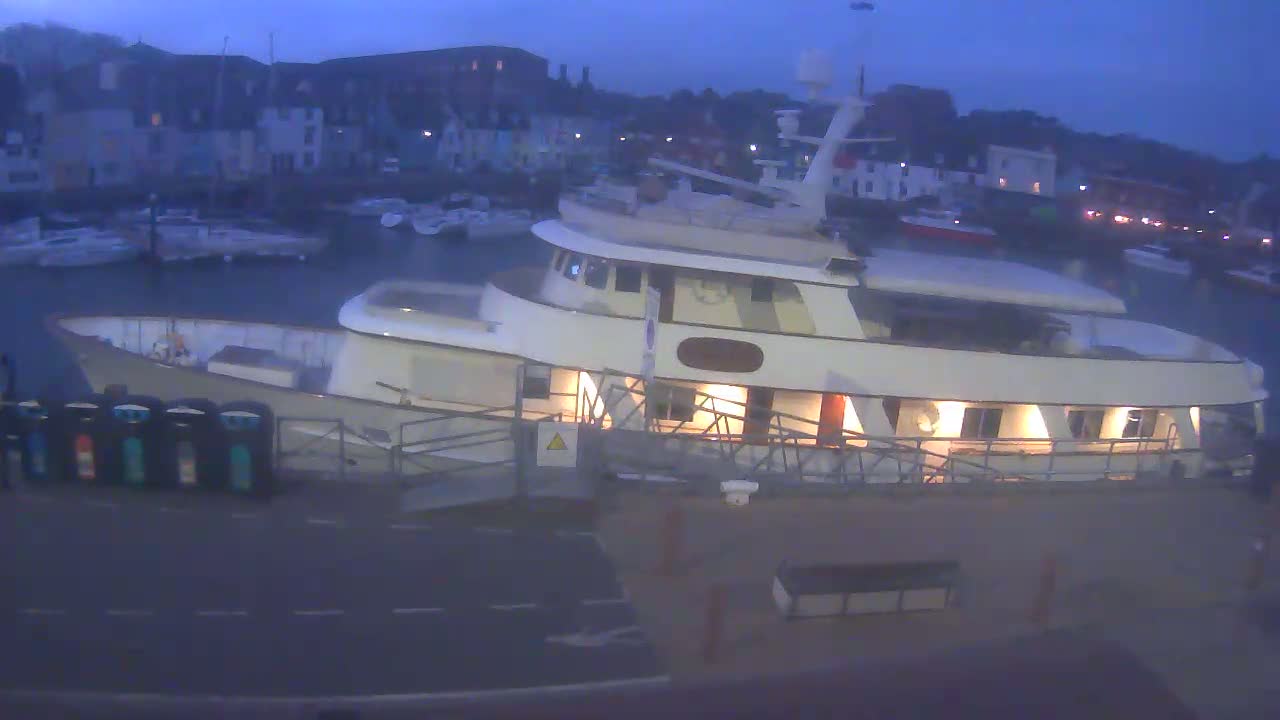 A large, lit-up yacht is docked at a harbor at twilight, with other boats visible in the background.