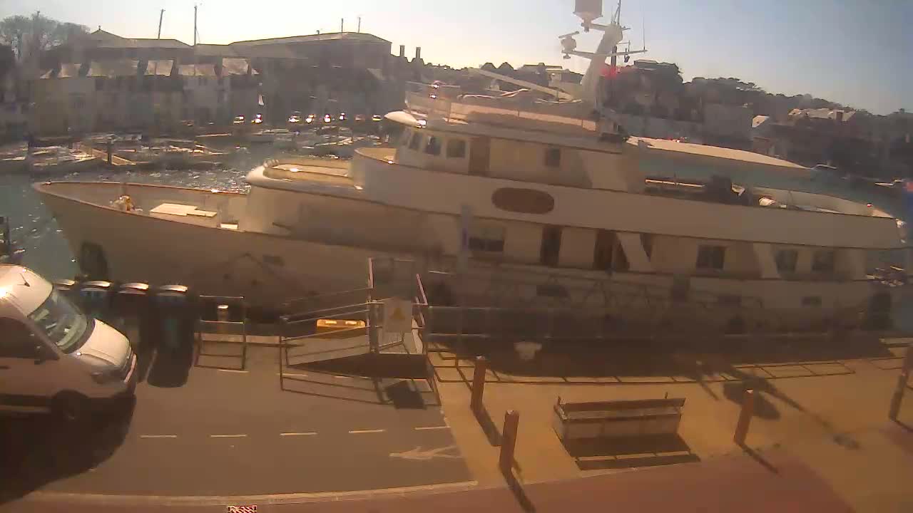 A large, light-colored motor yacht is docked at a harbor on a sunny day, with other boats visible in the background and a white van parked nearby.