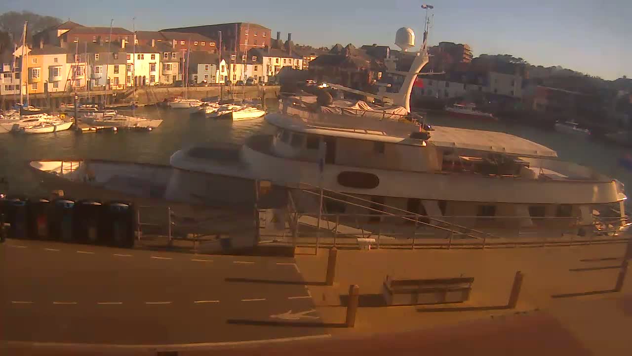 A large motor yacht is docked in a harbor filled with numerous smaller boats, under a clear, sunny sky.
