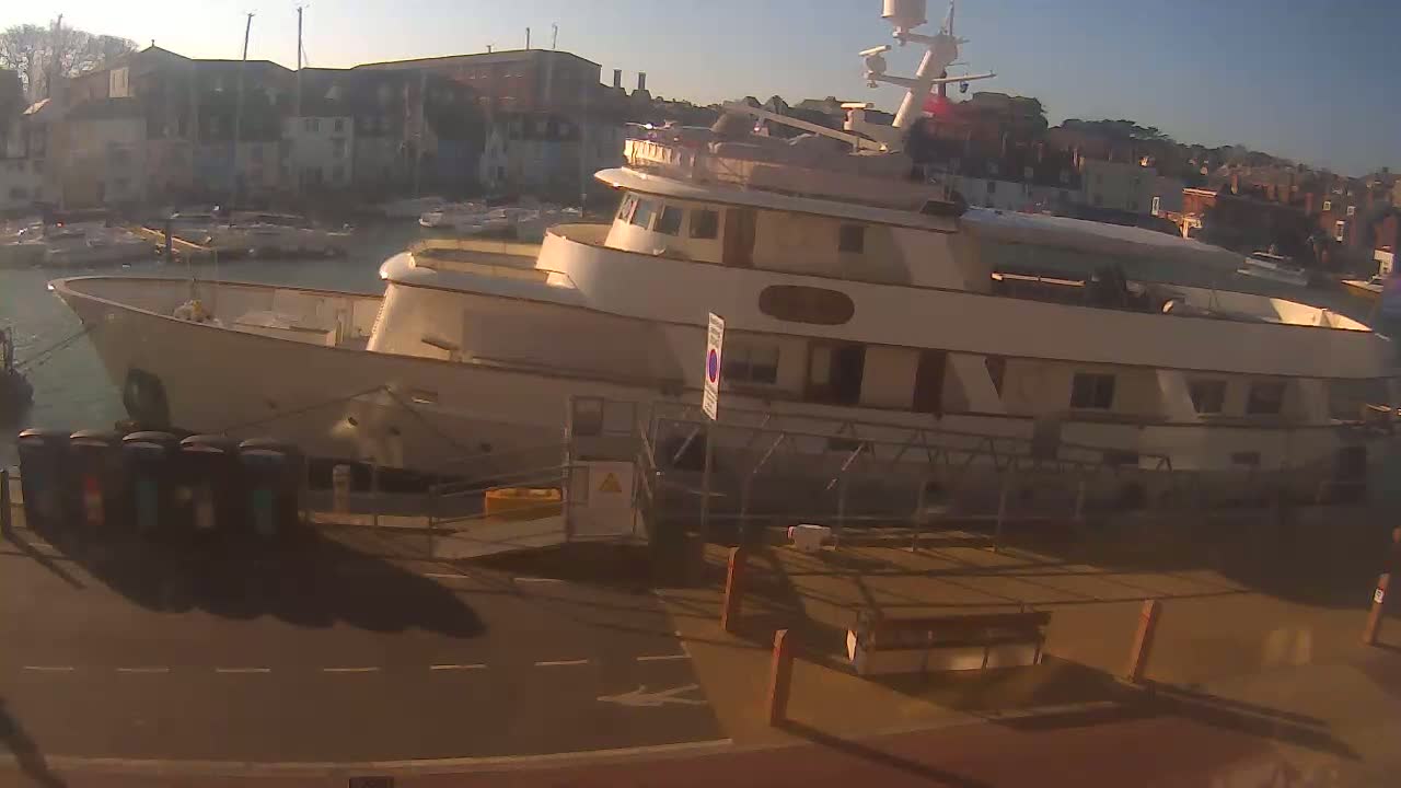 A large, light-colored yacht is docked at a harbor in the morning sun, with other smaller boats visible in the background.