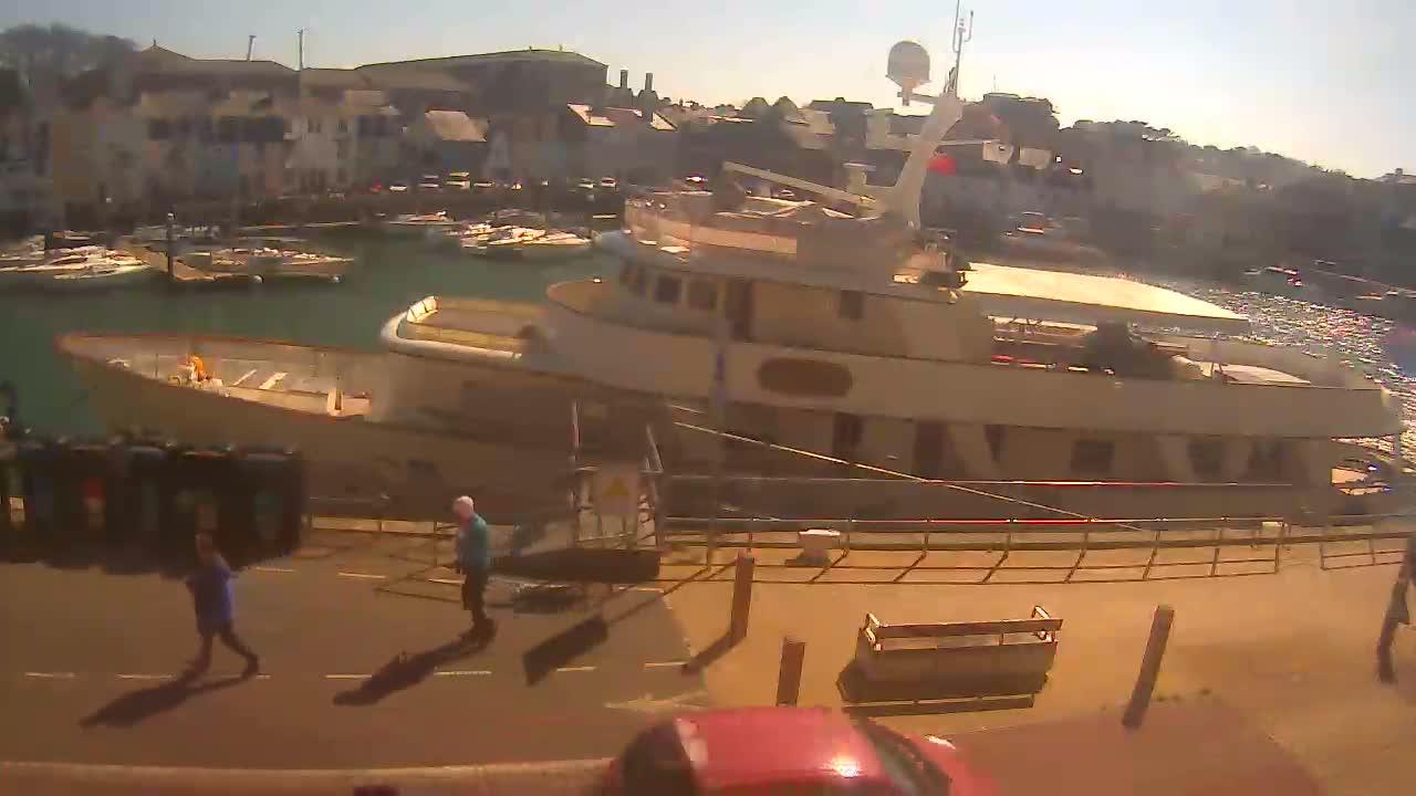 A large motor yacht is docked in a harbor on a sunny day, with several smaller boats moored nearby and people walking along a paved area.