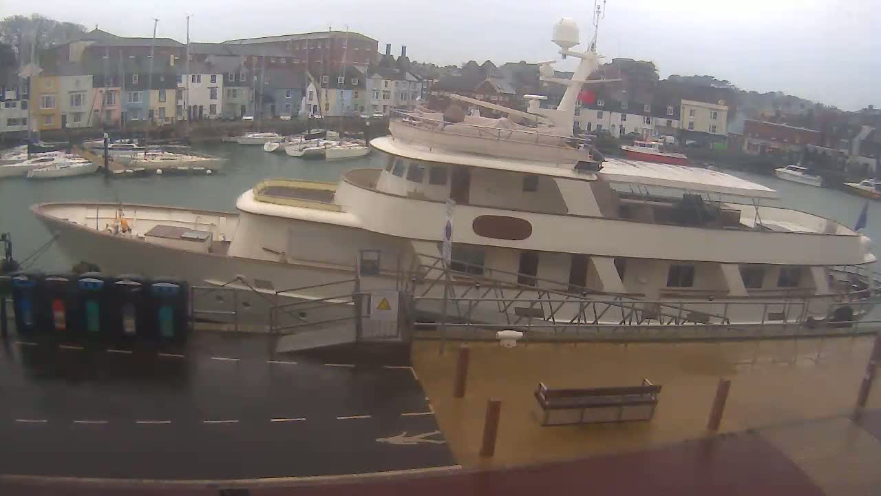 A large, cream-colored yacht is docked at a harbor on a cloudy day, with numerous smaller boats visible in the background.