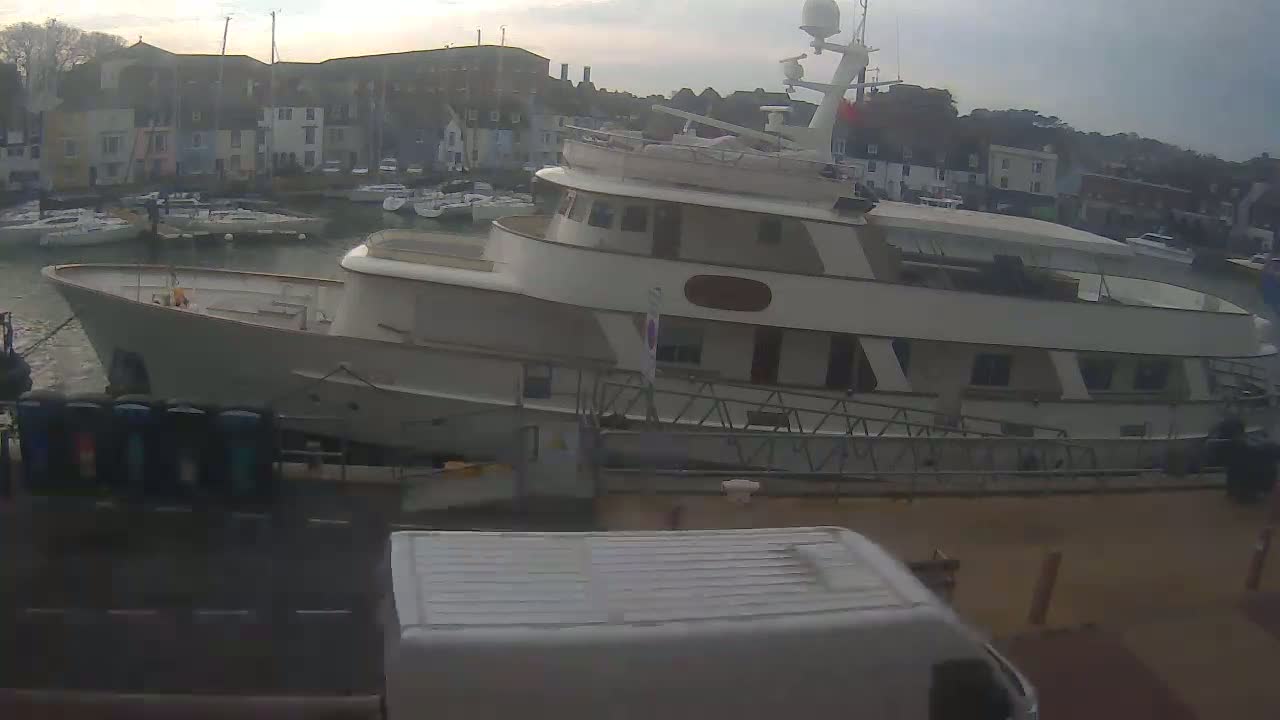 A large, light-colored yacht is docked in a harbor next to several smaller boats and buildings under an overcast sky.