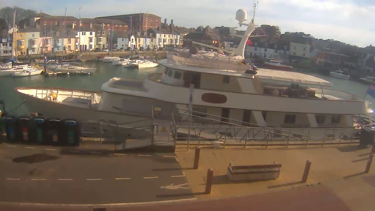 A large motor yacht is docked at a harbor on a sunny day, with numerous smaller sailboats moored in the background.