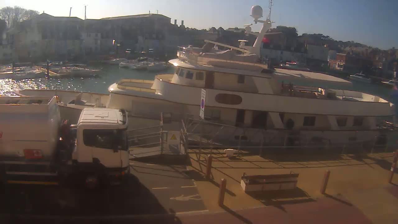A large, light-colored yacht is docked at a harbor on a sunny day, with a fuel tanker truck parked nearby.