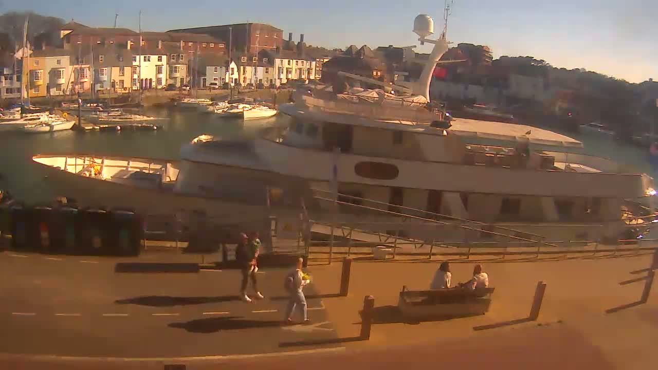 A large motor yacht is docked in a harbor filled with smaller boats, under sunny skies, with several people walking along the waterfront.