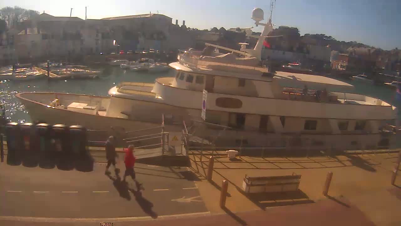 A large, light-colored yacht is docked at a harbor on a sunny day, with several smaller boats moored nearby and two people walking on the dock.