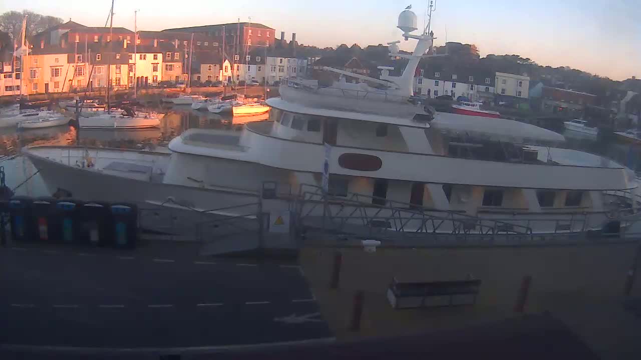 A large motor yacht is docked in a harbor at sunrise, with many smaller sailboats visible in the background.