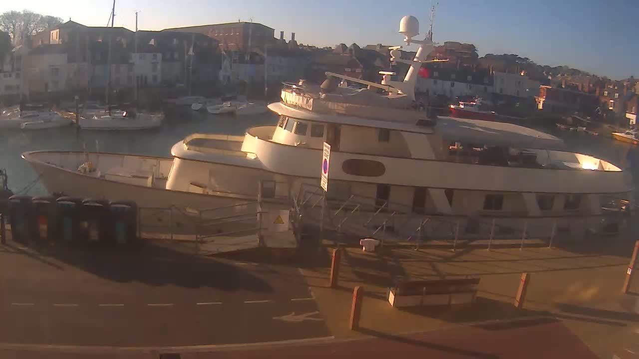 A large, light-colored motor yacht is docked at a marina on a sunny morning, with other boats visible in the background.