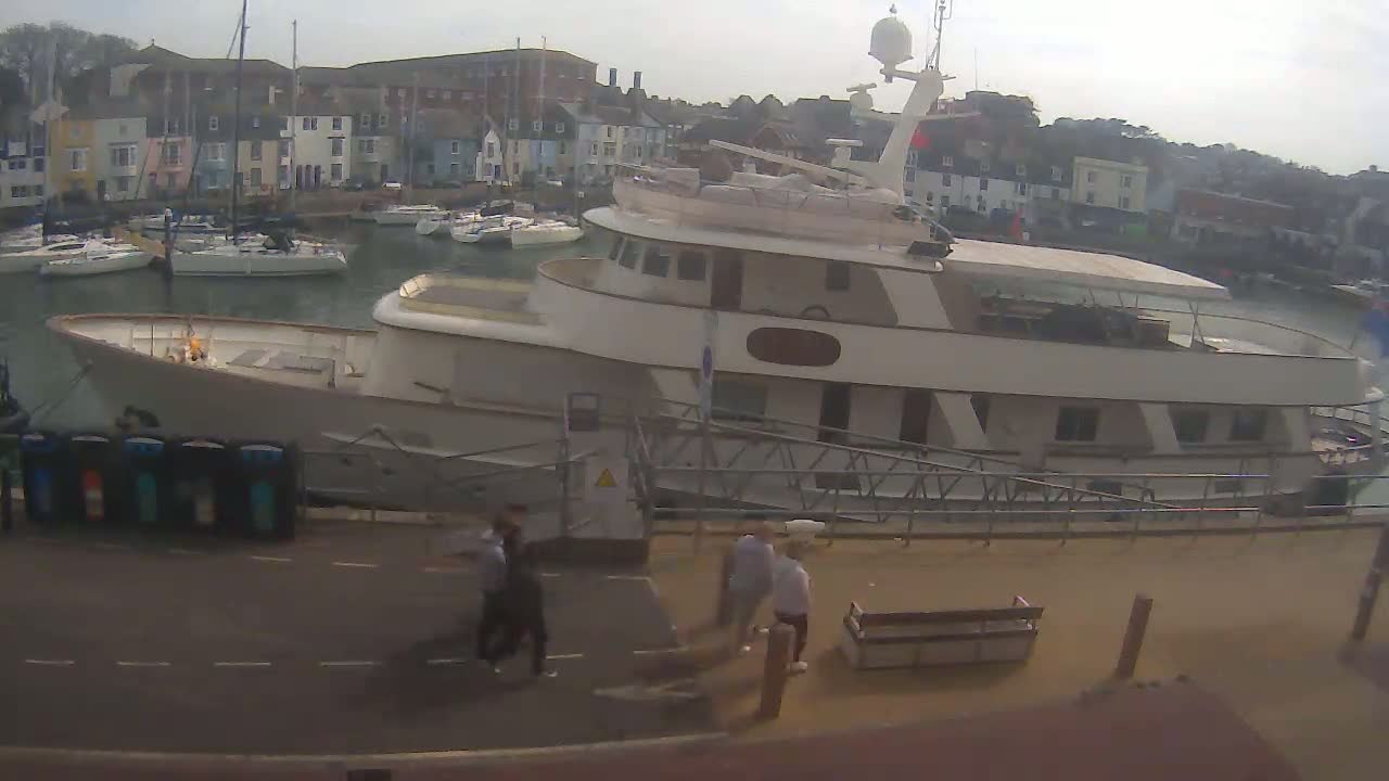 A large white motor yacht is docked at a marina on an overcast day, with several smaller sailboats visible in the background.