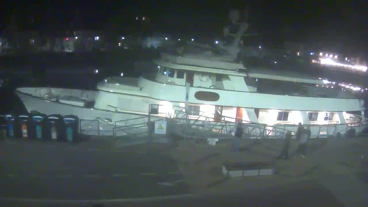 A large, white yacht is docked at night, with several people standing near a gangway leading to the vessel.