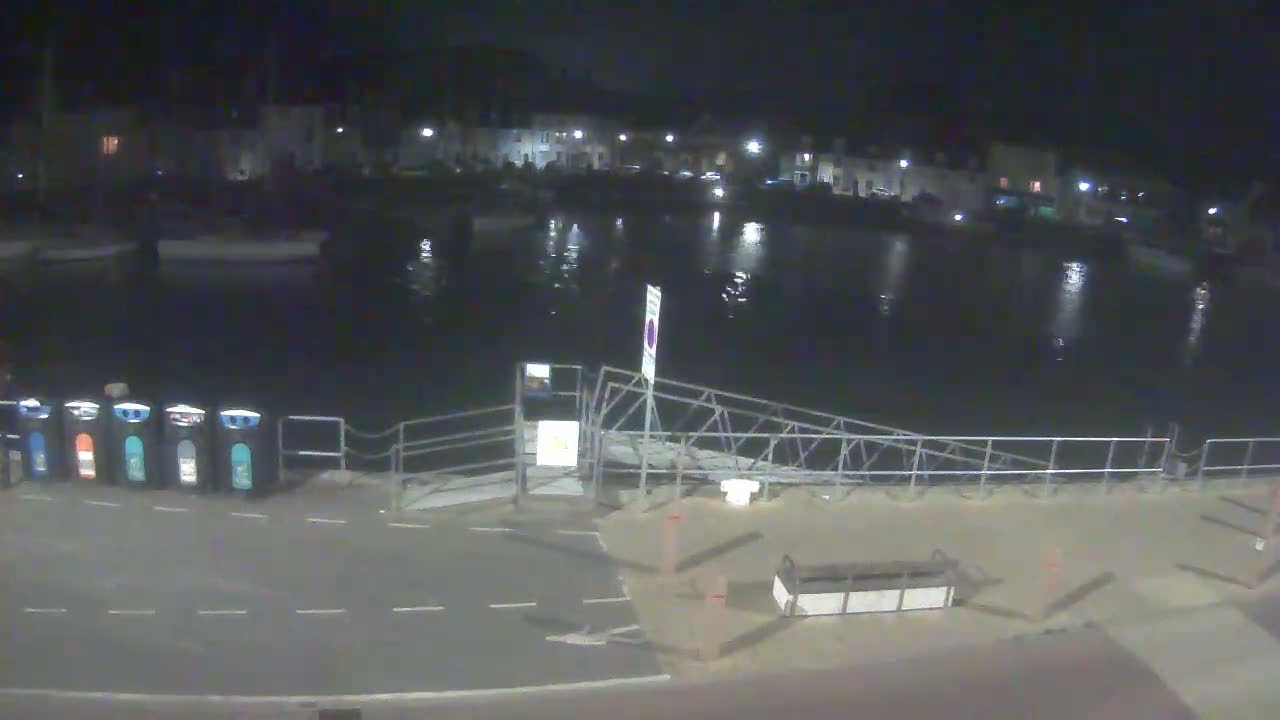 A nighttime view of a calm harbor with a metal gangway leading to a paved area with recycling bins, and buildings illuminated in the background.
