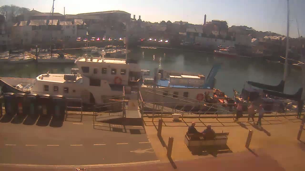 A sunny harbor scene shows several boats docked near a stone building, with people walking and sitting on a nearby quay.
