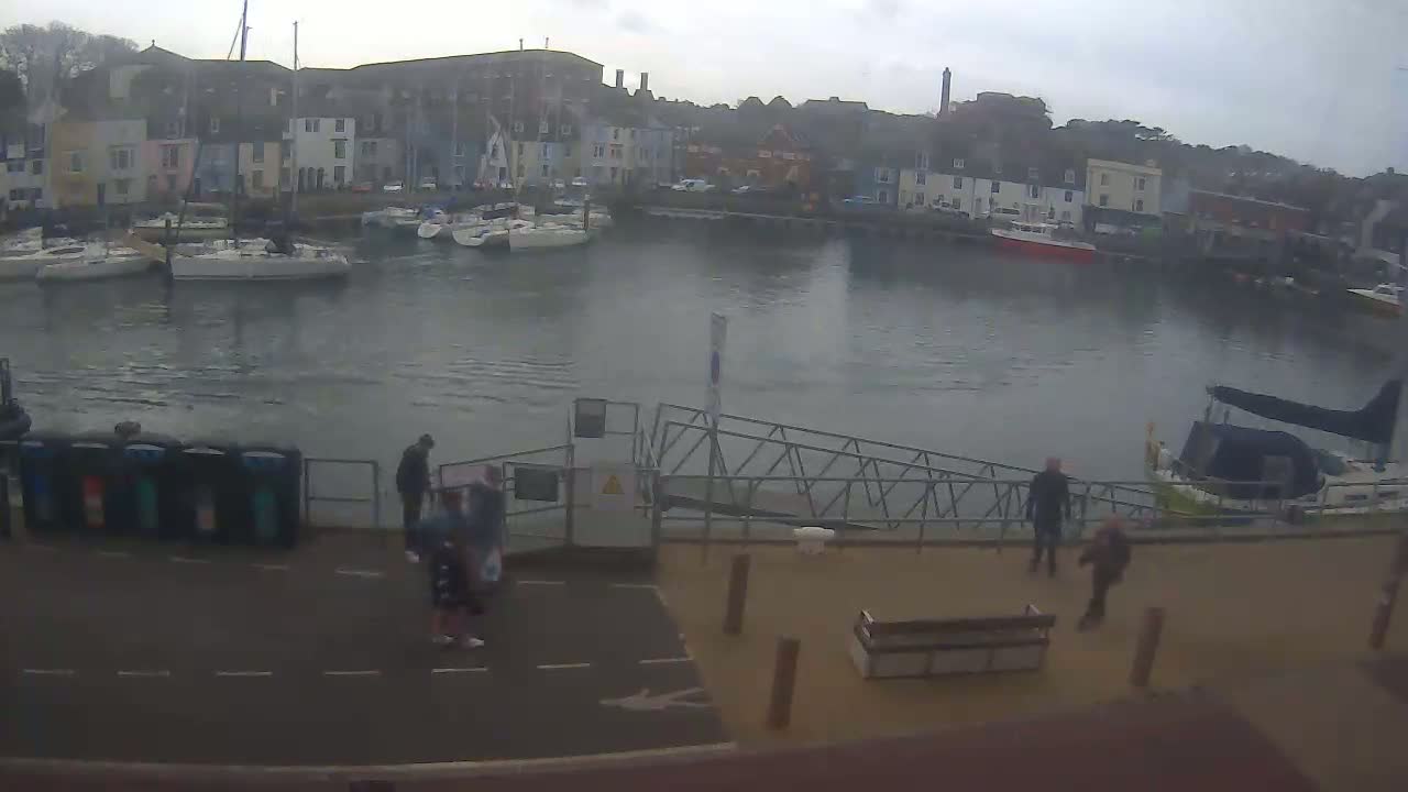 A harbor filled with sailboats under an overcast sky, with several people walking near a gangway and a bench.