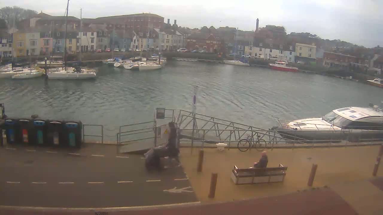 A harbor filled with sailboats and motorboats under an overcast sky, with people walking near a ramp and sitting on a bench.