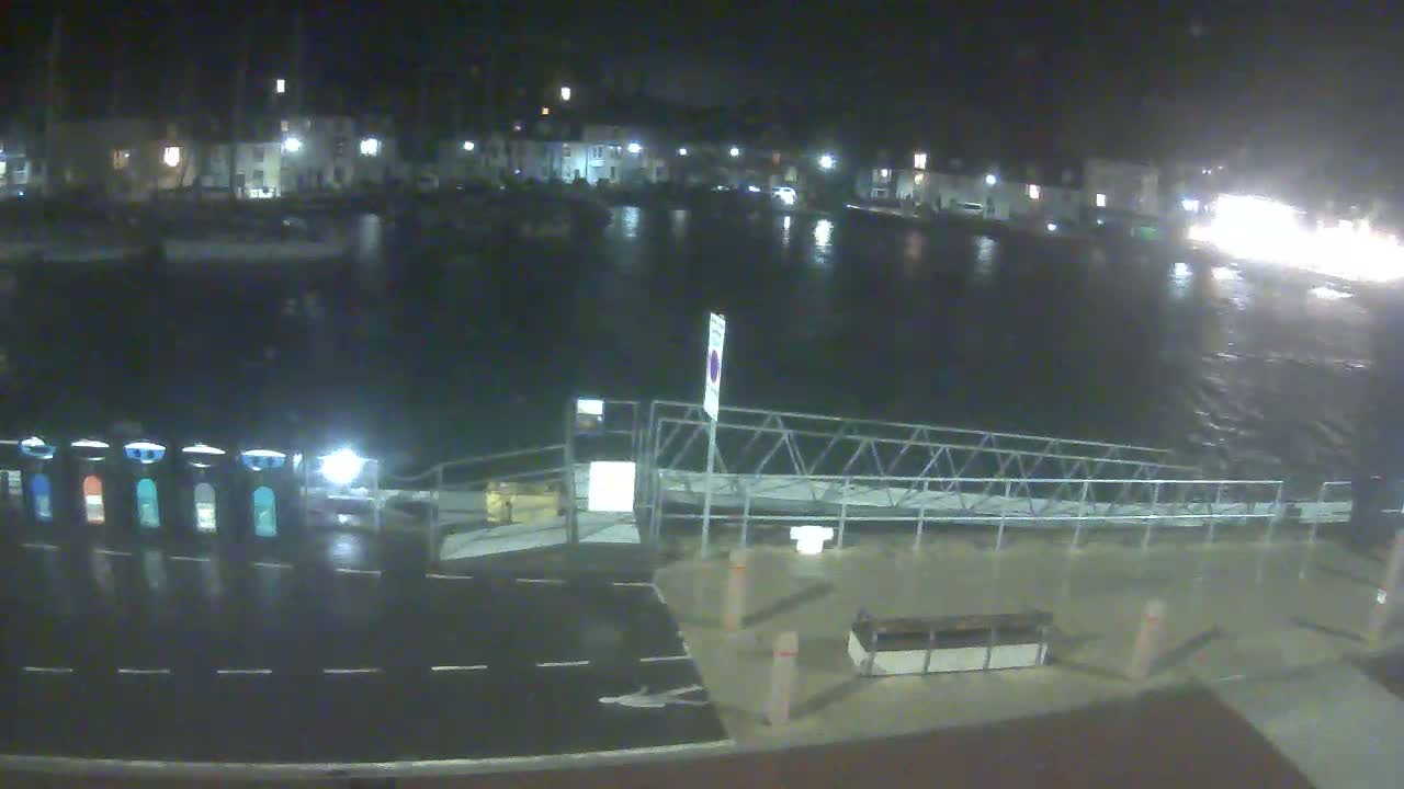 A nighttime view of a harbor with boats moored, a wet dockside area, and buildings with lights on across the water under dark, possibly rainy conditions.