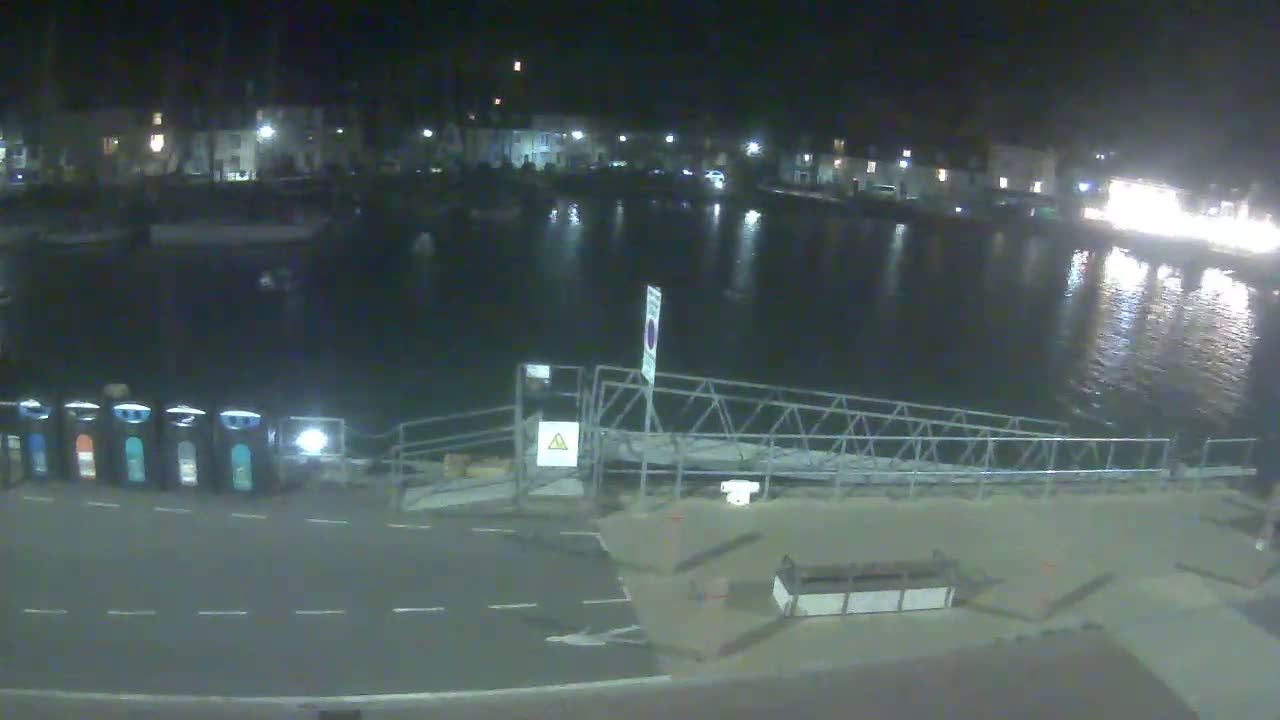 A nighttime view of a calm harbor with buildings lining the shore and a metal gangway leading to a paved area.