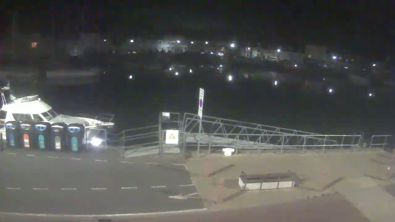 A white boat is docked at a harbor at night, next to a metal ramp, with several buildings faintly visible in the background under a dark sky.