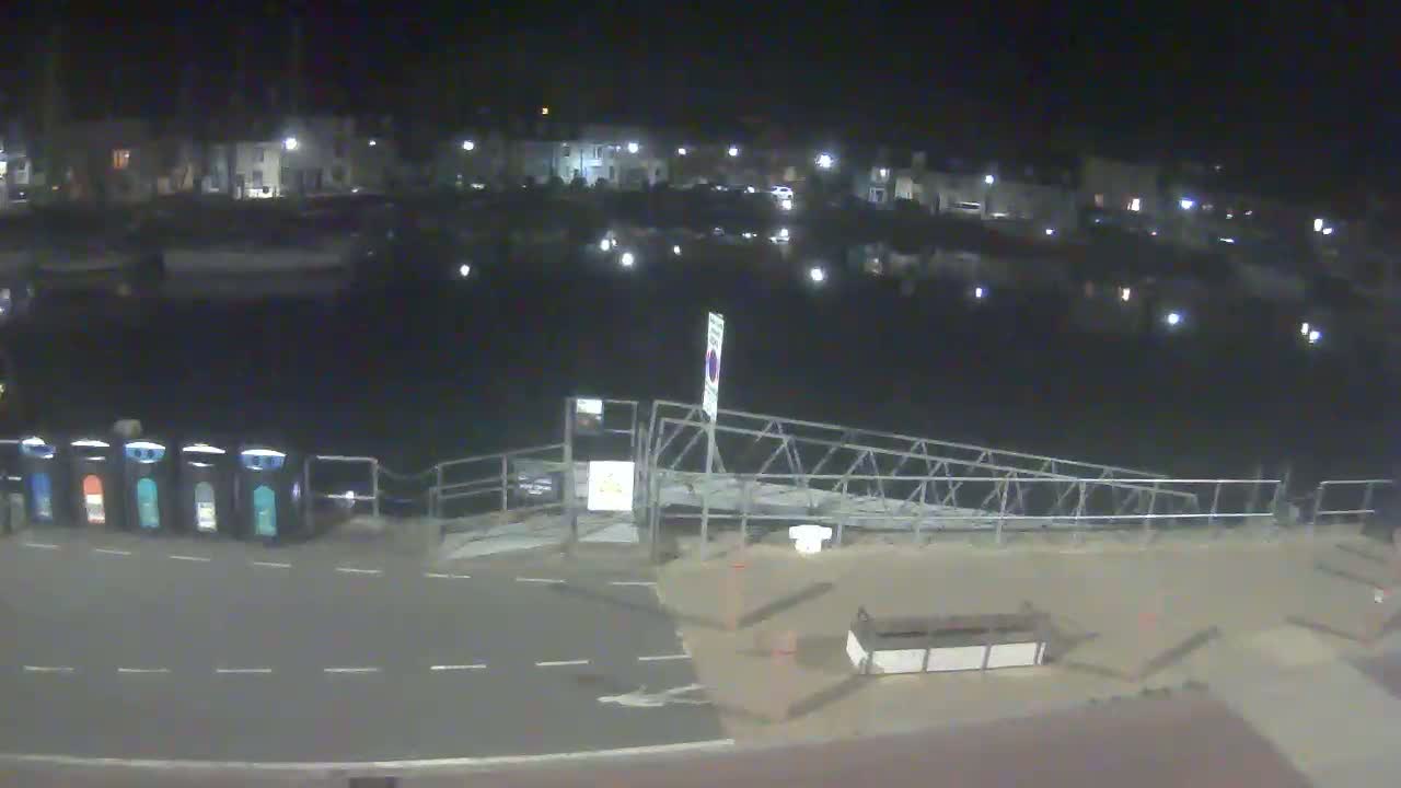 A nighttime view of a calm harbor with buildings across the water, a metal ramp leading to the water, and several recycling bins along a paved area.