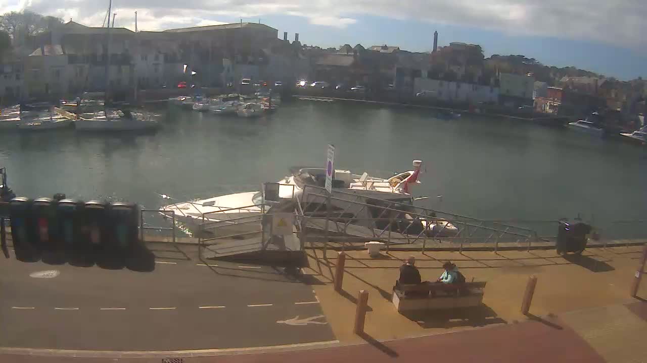 A harbor filled with boats under a partly cloudy sky, with a larger boat docked at a ramp where two people sit on a bench.