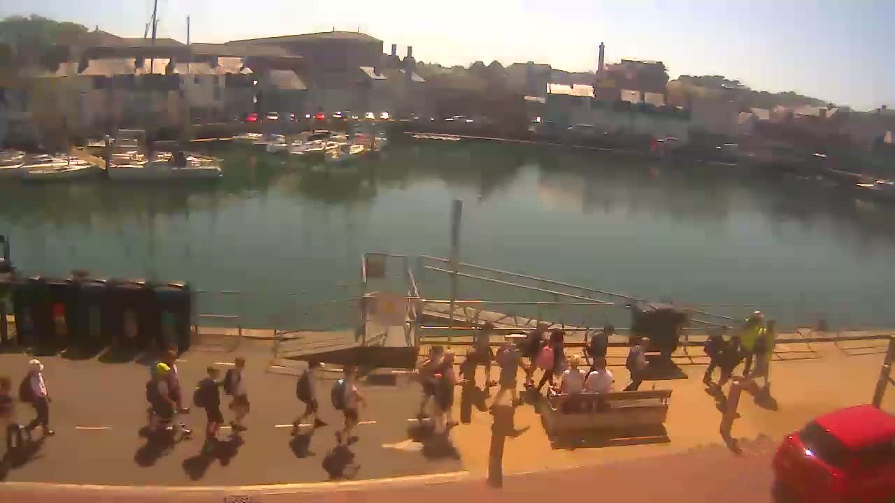 A sunny day at a harbor shows a calm body of water with moored boats, a group of people walking along a waterfront path, and a red car parked nearby.