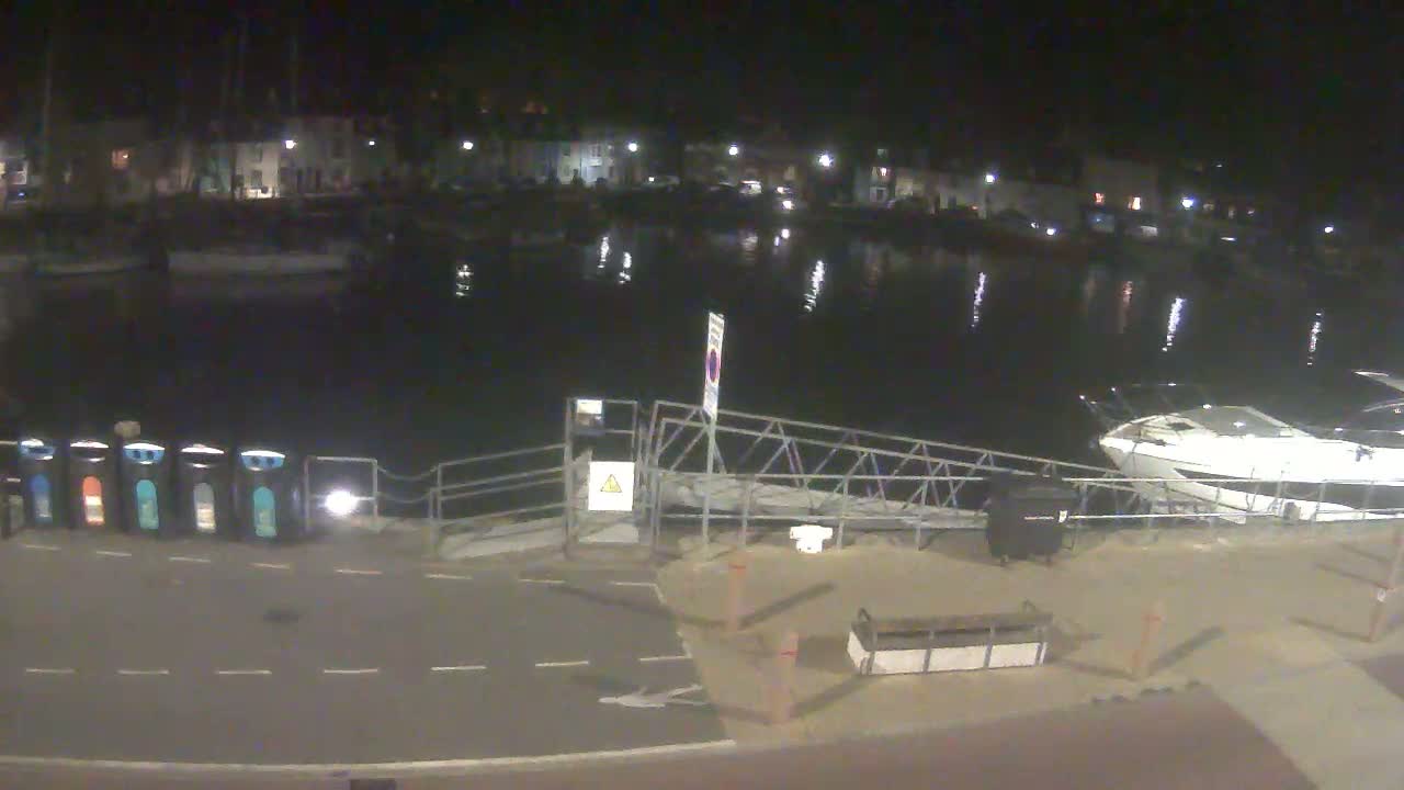 A nighttime view of a calm harbor with boats moored, a metal gangway, and several recycling bins on a paved area near waterfront buildings.