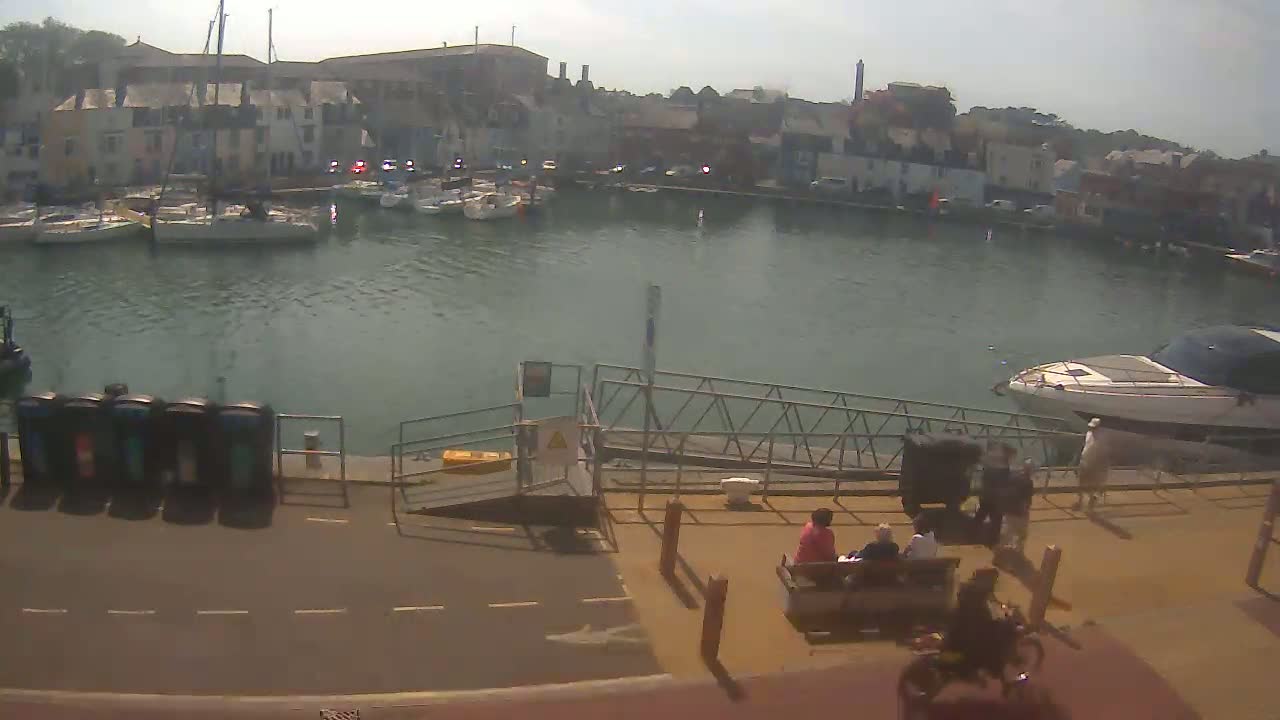 A harbor filled with sailboats and motorboats under a hazy, sunny sky, with people sitting on a bench and a cyclist riding by near a paved walkway.
