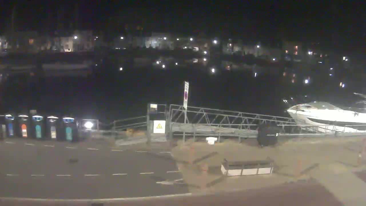 A nighttime view of a marina with boats docked, a metal walkway, and several waste bins along a paved area.
