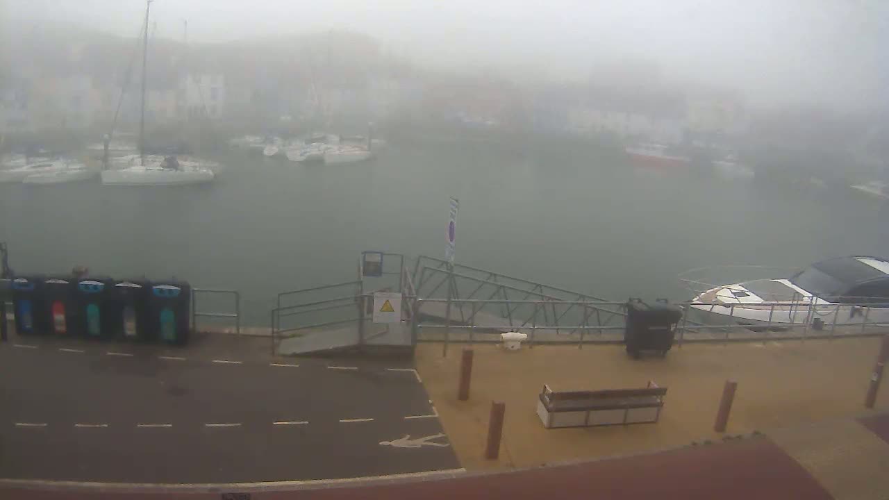 A foggy harbor scene shows several boats moored at a dock with a ramp and a bench on the shore.